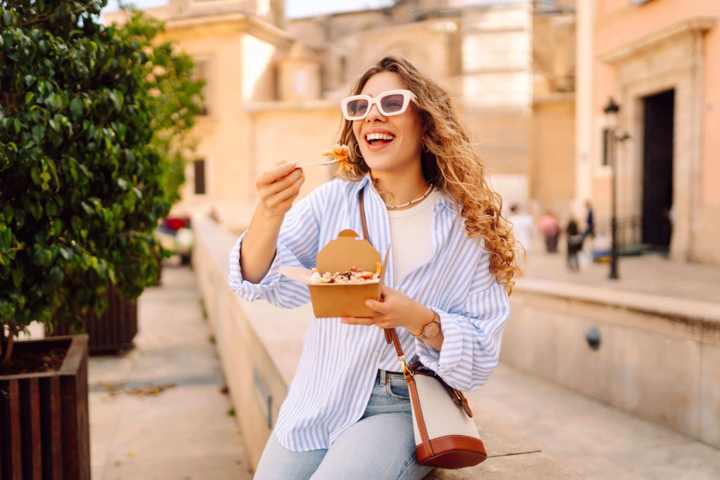 Lady eating food from a street-food style box wearing sunglasses. She is happy.