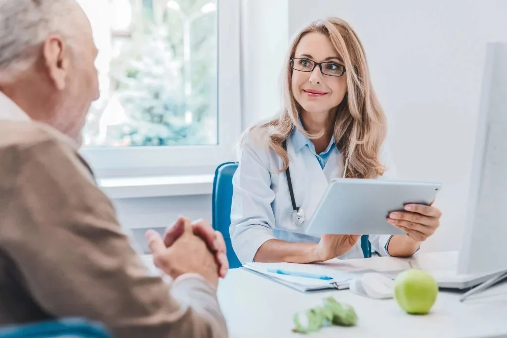 A smiling nutritional expert with long blond hair, looking in the direction of her elderly customer as she types information on a tablet.