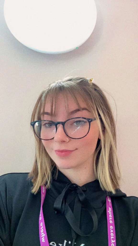 young woman with round glasses taking a selfie in a black top with a pink lanyard around her neck