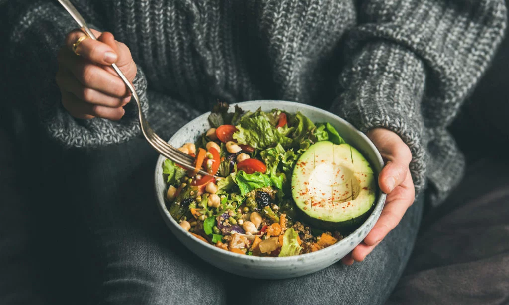woman in a grey sweater and grey jeans eating an avocado salad