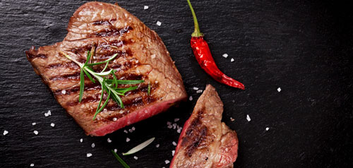 a slab of grilled beef cut in two pieces with a red chili to the side and sprinkled with salt against a black background