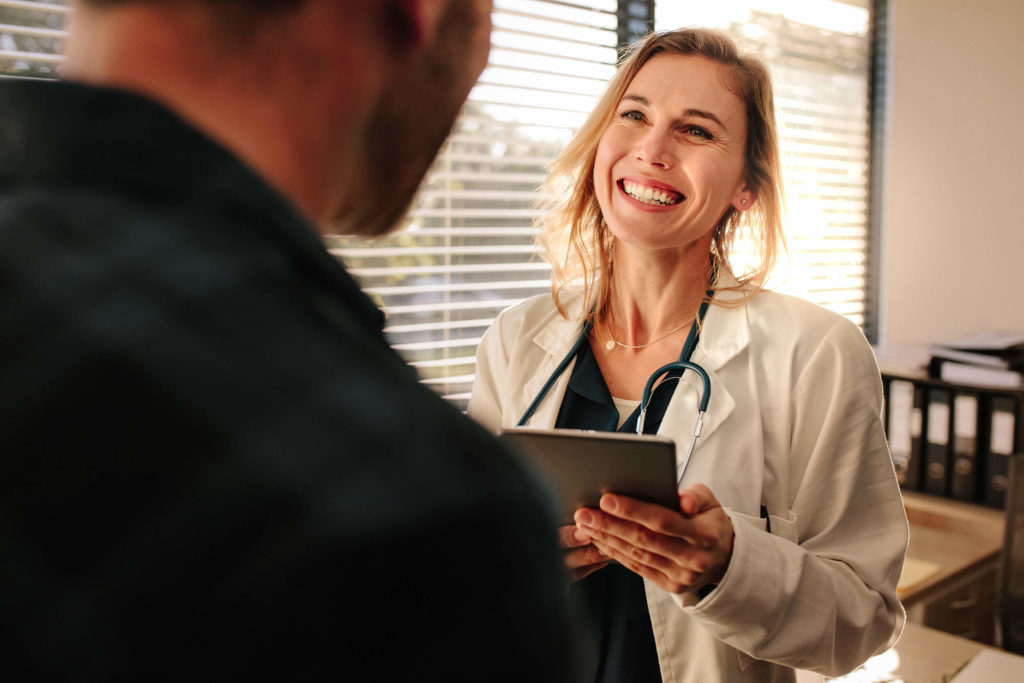Woman in a sunlit office smiling to a customer while holding a tablet in her hands and a stethoscope around her neck