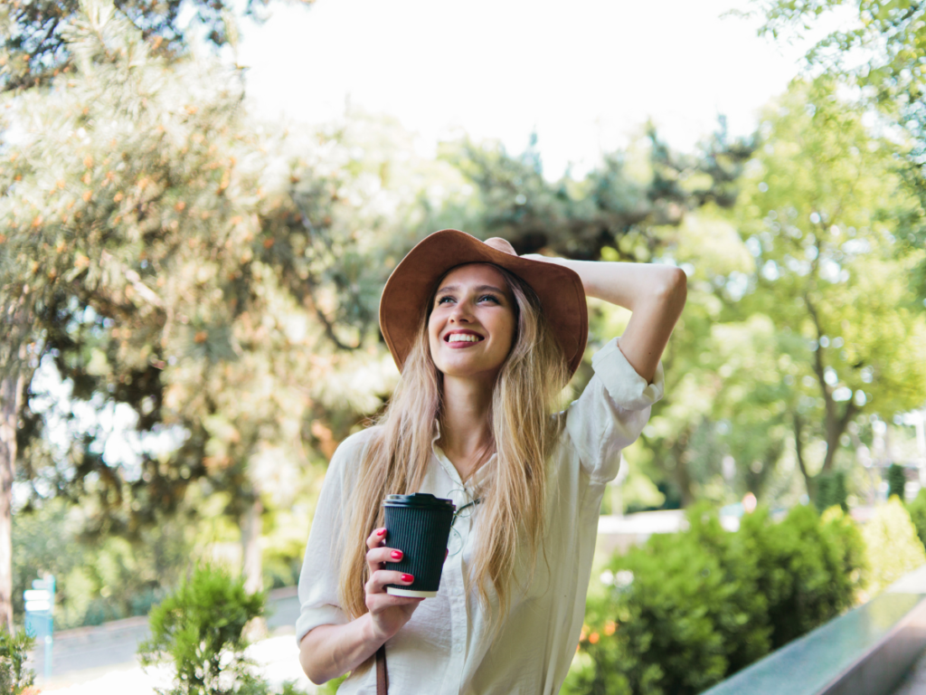 Young woman smiling in a park, holding a cup of coffee in one hand and holding on to a brown hat sat on her head.