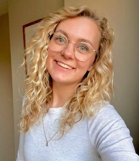 A young woman with glasses, blond curls and a white top posing for the camera.