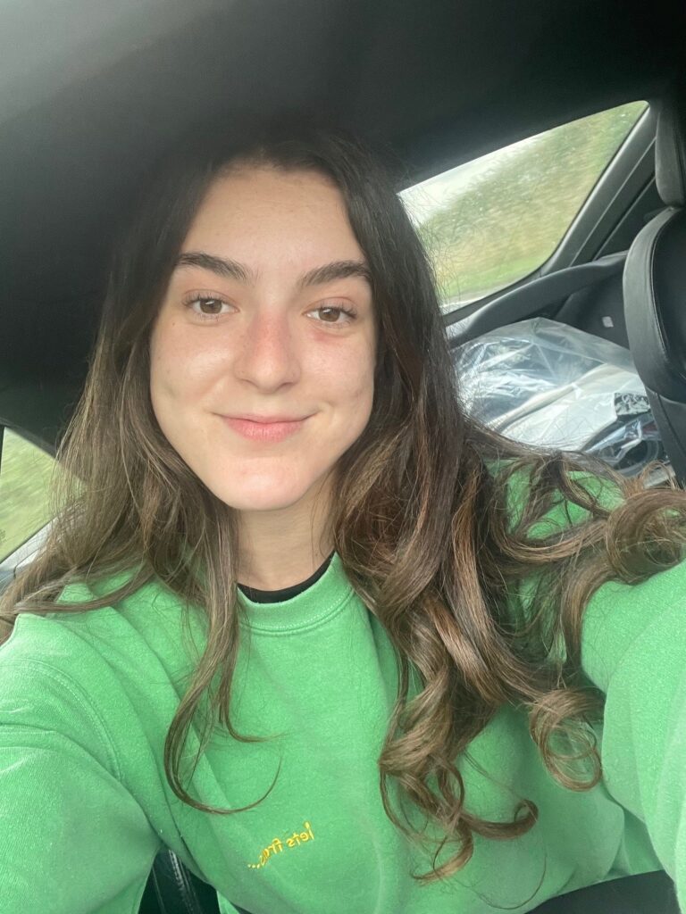 young woman with long brown hair posing for a selfie in a car and wearing a green sweater