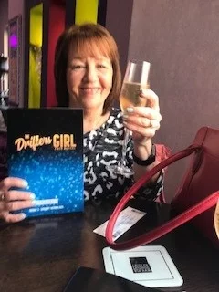 middle aged woman posing with a glass of beer and a book in her hands while smiling at the camera