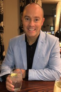 Smartly dressed man in light blue suit smiling at the camera and holding a drink in his hand