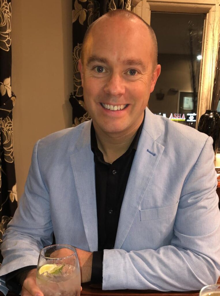 Smartly dressed man in light blue suit smiling at the camera and holding a drink in his hand