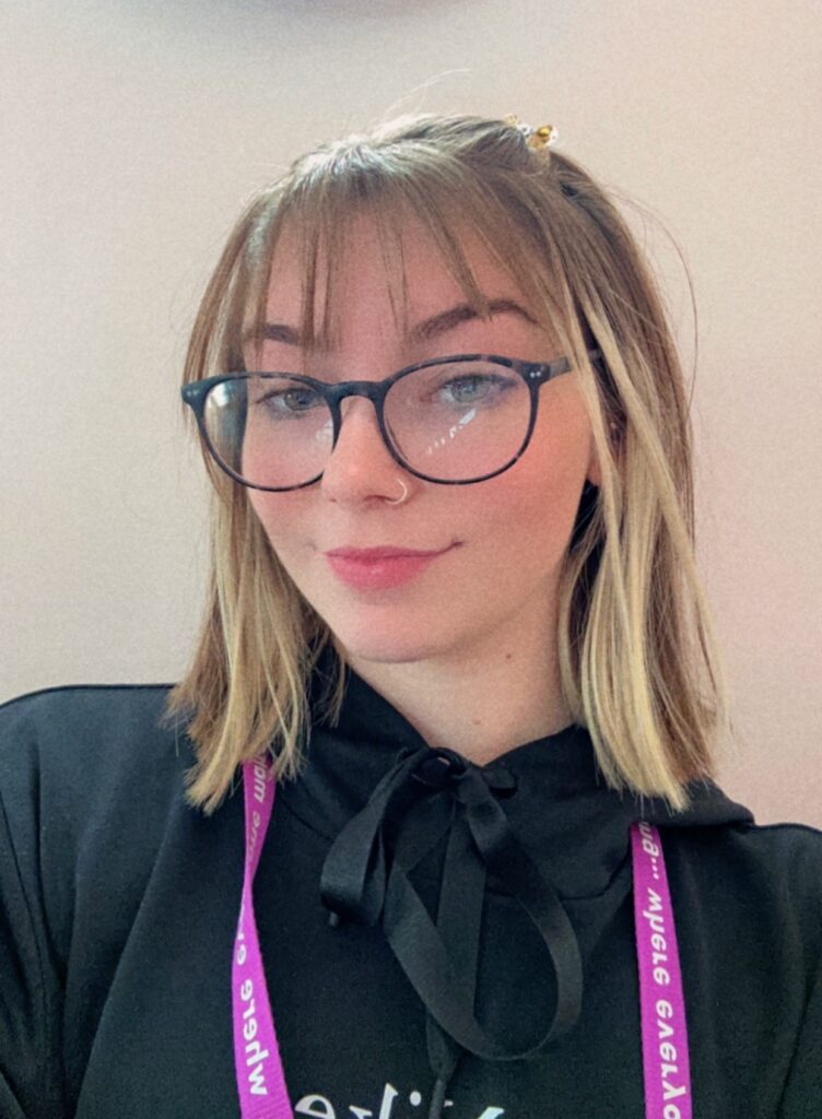 young woman with round glasses taking a selfie in a black top with a pink lanyard around her neck